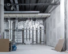 Ein großer, hoher Raum mit Betonoptik-Wänden, im Baustellenzustand. Im Hintergrund sind dick isolierte Rohre an der Wand, die miteinander verbunden sind. Schwarze Decke mit Kabelschächten.