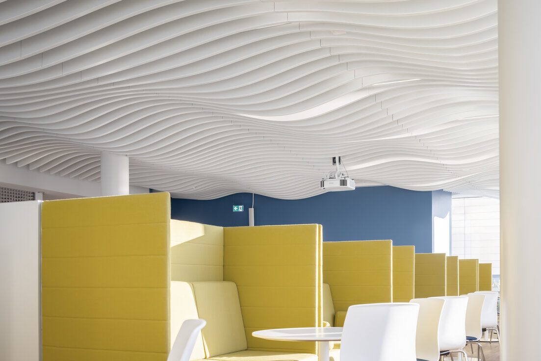 Moderne Kantine im Bürogebäude mit markanter Lamellendecke von Lindner und gelben Sitznischen für eine inspirierende Pausenatmosphäre.