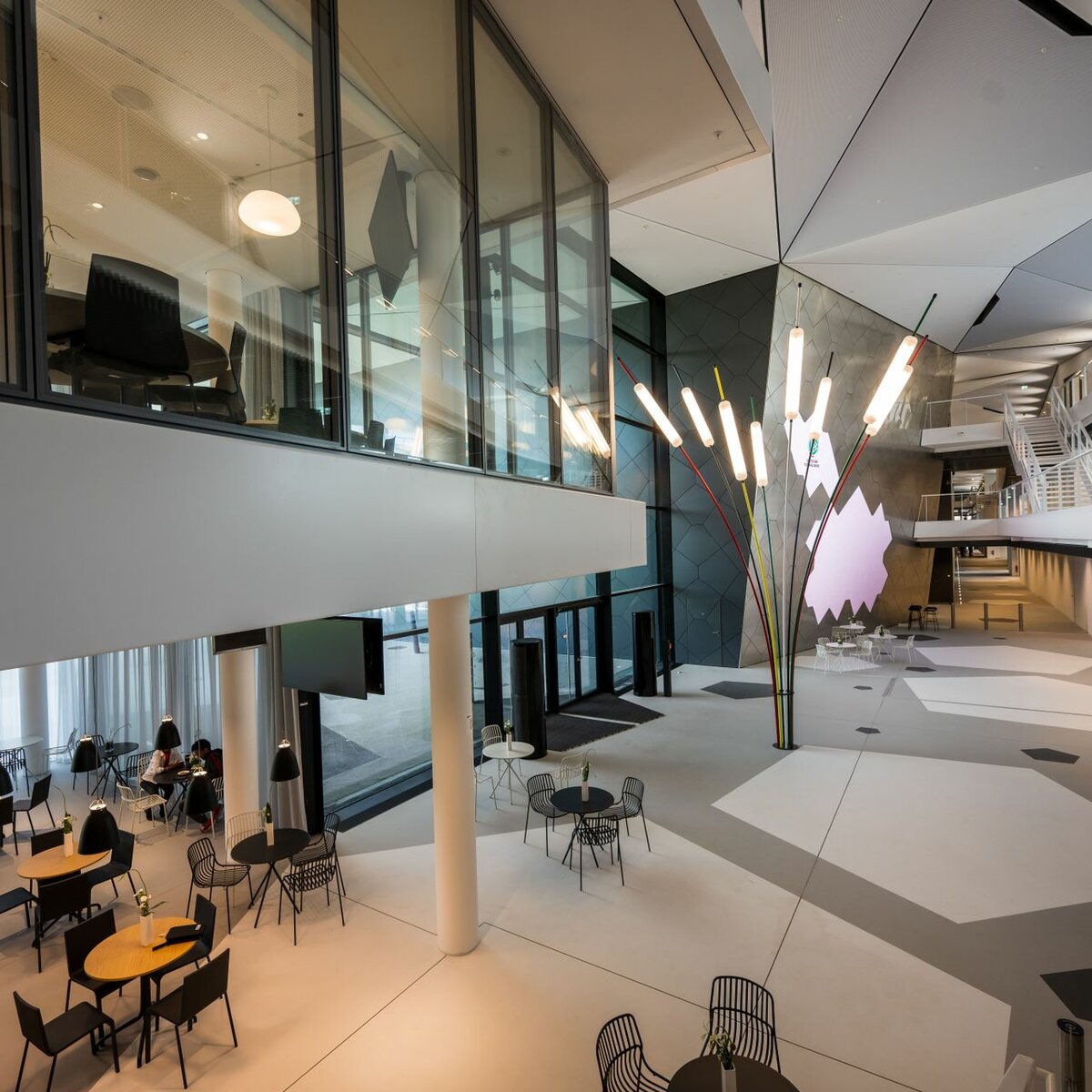 Eingangsbereich der DFB-Akademie in Frankfurt mit modernen Sitzgruppen im Erdgeschoss. Im Foyer stehen mehrere Tische und Stühle in hellen, offenen Bereichen. Im ersten Obergeschoss sind die verglasten Trennwände von Lindner zu sehen, die den Raum transparent und lichtdurchflutet wirken lassen.