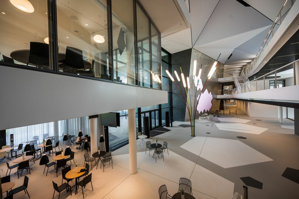 Eingangsbereich der DFB-Akademie in Frankfurt mit modernen Sitzgruppen im Erdgeschoss. Im Foyer stehen mehrere Tische und Stühle in hellen, offenen Bereichen. Im ersten Obergeschoss sind die verglasten Trennwände von Lindner zu sehen, die den Raum transparent und lichtdurchflutet wirken lassen.