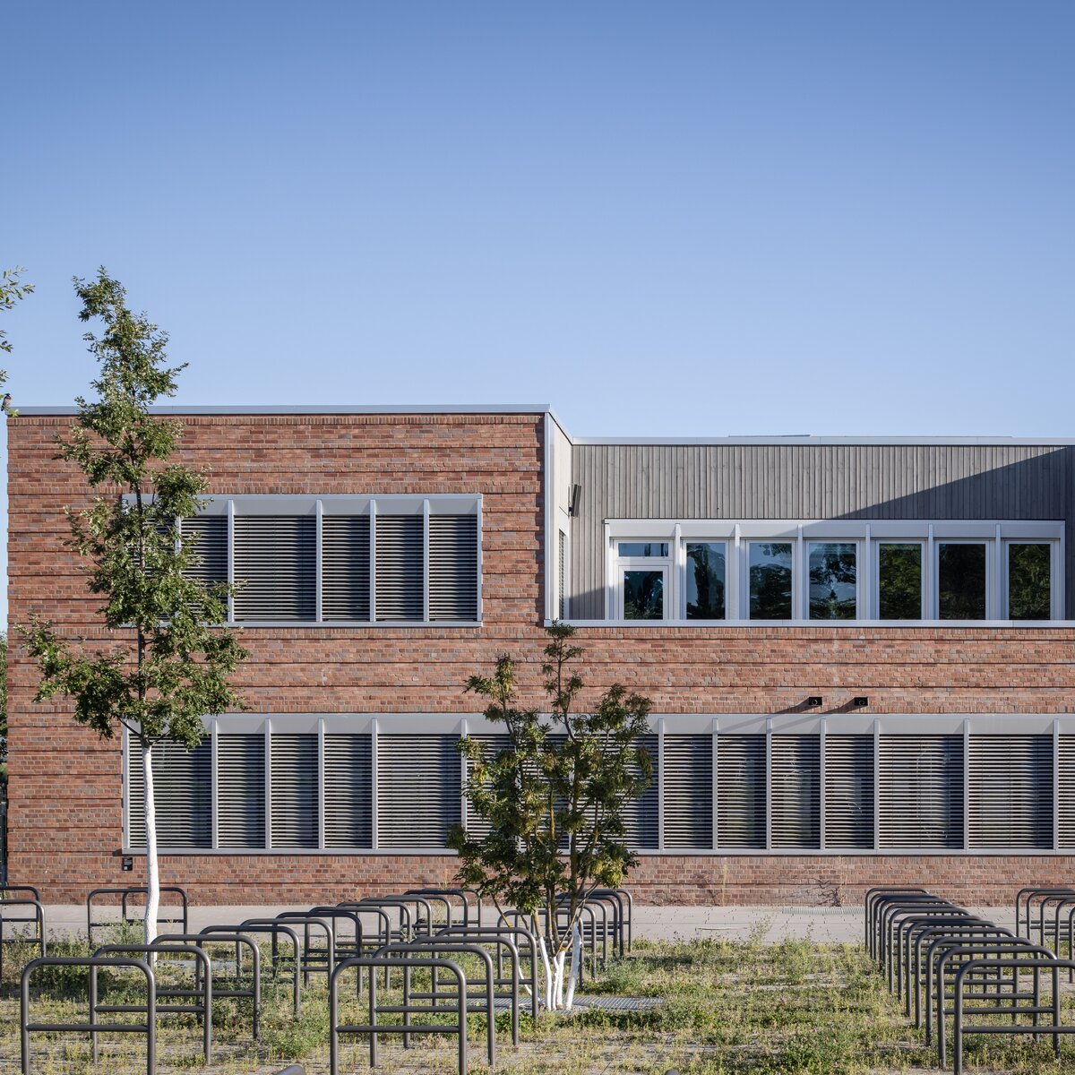 Außenansicht der Clay Schule in Berlin-Neukölln, Neubau mit rostbrauner Fassade, weißen Fenstern, dunklen Lamellen und grüner Fläche mit Bäumen im Vordergrund. | © © www.Lindner-Group.com