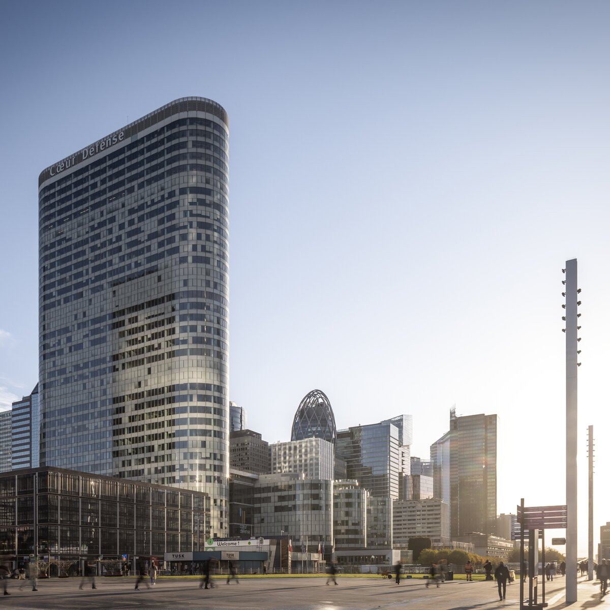 Außenansicht des Bürokomplexes Cœur Défense in Paris mit Vorplatz, umliegenden Hochhäusern, Passanten und tief stehender Sonne | © © www.Lindner-Group.com